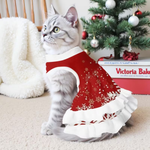 A gray tabby cat wearing a red and white Christmas-themed pet sweater dress with white snowflake patterns, sitting on a beige carpet in front of a decorated Christmas tree and a white blanket.