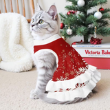 A gray tabby cat wearing a red and white Christmas-themed pet sweater dress with white snowflake patterns, sitting on a beige carpet in front of a decorated Christmas tree and a white blanket.