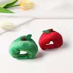 Two plush apple-shaped headbands, one green and one red, displayed on a clean white surface with decorative tulips in the background.