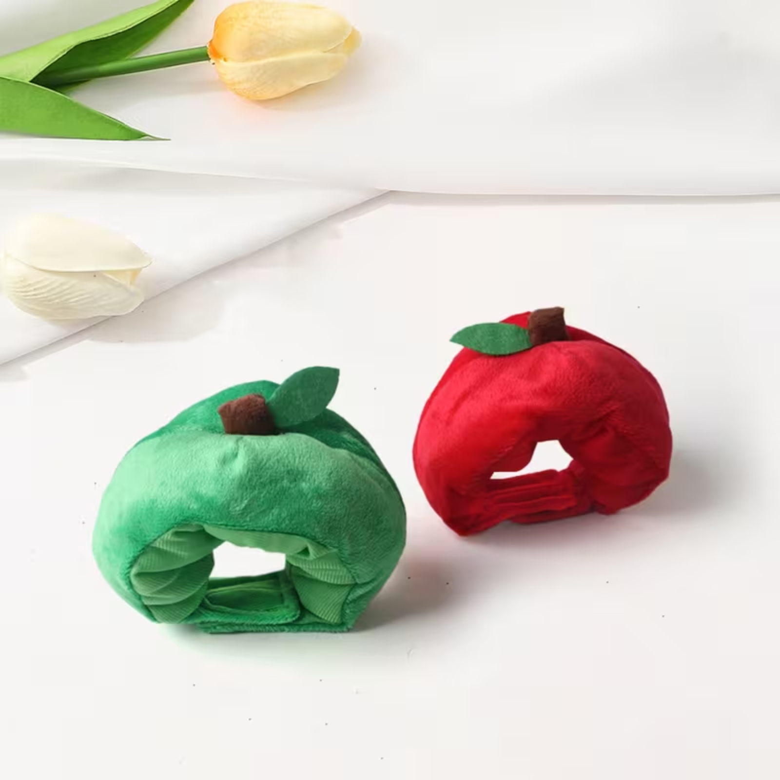 Two plush apple-shaped headbands, one green and one red, displayed on a clean white surface with decorative tulips in the background.