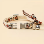 A floral-patterned pet collar with a matching fabric bow, gray plastic buckle, and a small silver bell, displayed on a solid light beige background.