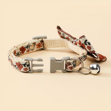 A floral-patterned pet collar with a matching fabric bow, gray plastic buckle, and a small silver bell, displayed on a solid light beige background.