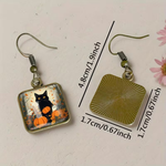 Square dangle earrings shown on a light beige surface with measurement lines and text indicating dimensions. One earring features a black cat with orange pumpkins in a fall-themed glass panel, and the other shows the back of the earring with a bronze-toned sunburst square charm.