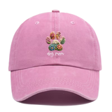 A light pink baseball cap featuring a colorful embroidery-style dog paw print with flowers and the words 'dog mom' in white script on the front. 