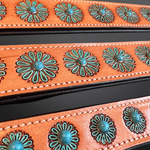 Close-up of a western-style leather belt strap in a warm orange-brown tone, decorated with repeating turquoise-blue floral studs and dark edging.