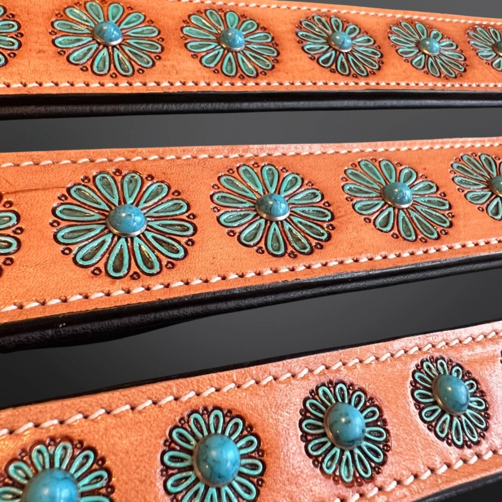 Close-up of a western-style leather belt strap in a warm orange-brown tone, decorated with repeating turquoise-blue floral studs and dark edging.