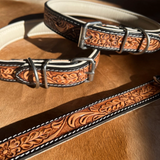 A close-up of a tooled leather belt with floral embossing, silver-tone metal buckle and hardware, and white contrast stitching, laid on a wooden surface.