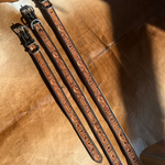 Three tooled leather straps with dark brown floral engravings and silver-tone buckles, laid diagonally on a textured tan leather surface.