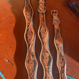 Three tooled leather straps laid flat on a wooden surface, featuring intricate floral carving and silver-toned metal hardware.