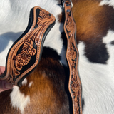 A close-up of a hand holding a tooled leather strap or bridle component against a cowhide background. The leather is tan and dark brown with intricate floral tooling and stitched edges.