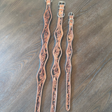 Three hand-tooled leather straps with silver metal buckles, laid out on a dark wood surface. The straps feature intricate floral and scrollwork carvings.
