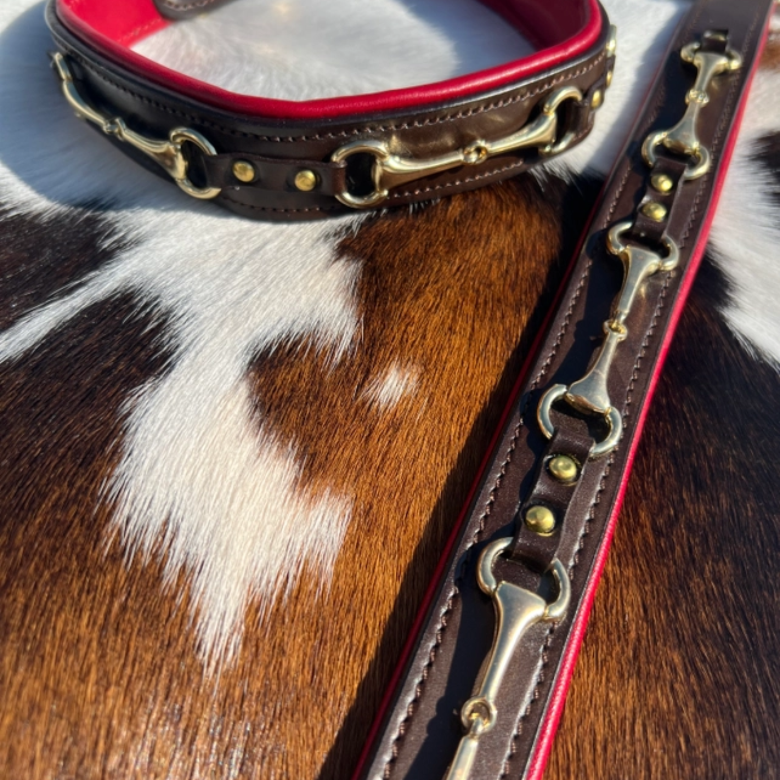 A close-up of a dark brown leather horsebit collar with red lining and brass hardware, laid on a brown and white cowhide surface.
