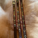 Three brown leather horse bits dog collars with gold-toned metal hardware laid out on a tan leather surface.