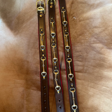 Three brown leather horse bits dog collars with gold-toned metal hardware laid out on a tan leather surface.
