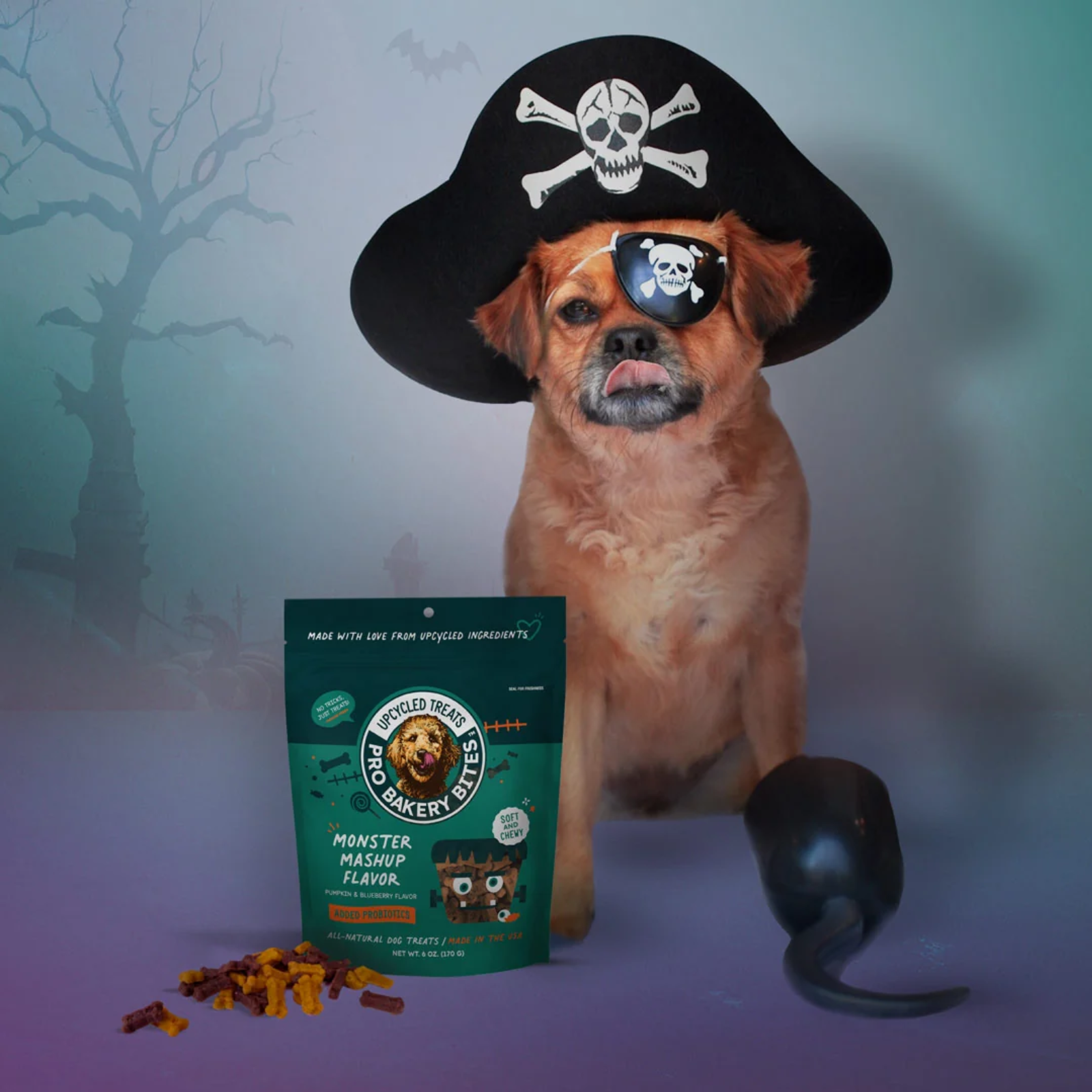 A small tan dog dressed as a pirate sits in front of a teal bag of 'Monster Mash' flavored treats, with scattered treats on a purple surface and a black toy hand hook beside it. The background is a misty blue-gray Halloween scene with silhouetted trees.