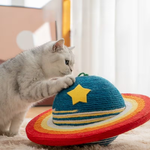 A silver tabby cat is pawing at a colorful woven cat toy shaped like a Saturn-like ringed planet, with a blue top, yellow star, and red, orange, and yellow bands.