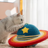 A silver tabby cat is pawing at a colorful woven cat toy shaped like a Saturn-like ringed planet, with a blue top, yellow star, and red, orange, and yellow bands.