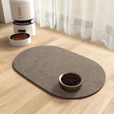 An oval brown pet mat on a light wood floor, with a metal bowl filled with kibble placed on top. A white automatic pet feeder and bowl are visible in the background near sheer curtains.