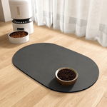 An oval-shaped black non-slip pet mat on a light wood floor, with a wooden bowl of dog food placed on top. In the background, a white automatic pet feeder and another food bowl are visible near sheer white curtains.