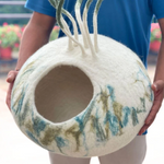 A cream, blue, and green felt pet cave with decorative strings on the top.